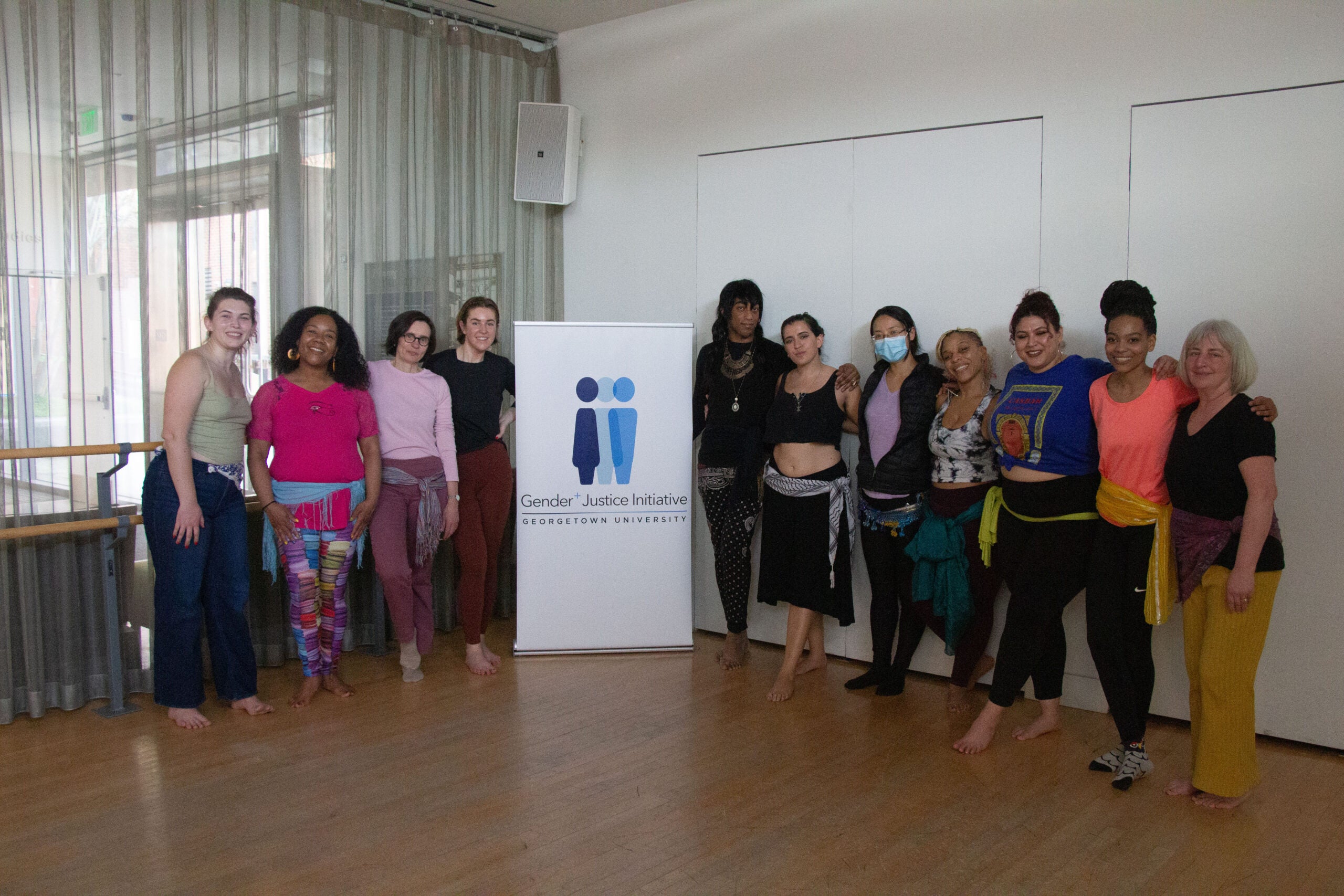 Eleven dancers pose for a photo in a dance studio. A roll up banner stands between the dancers. Four to the left of the banner and seven to the right. The dancers are of different races, ethnicities, and genders. Warda is the third one on the right.
