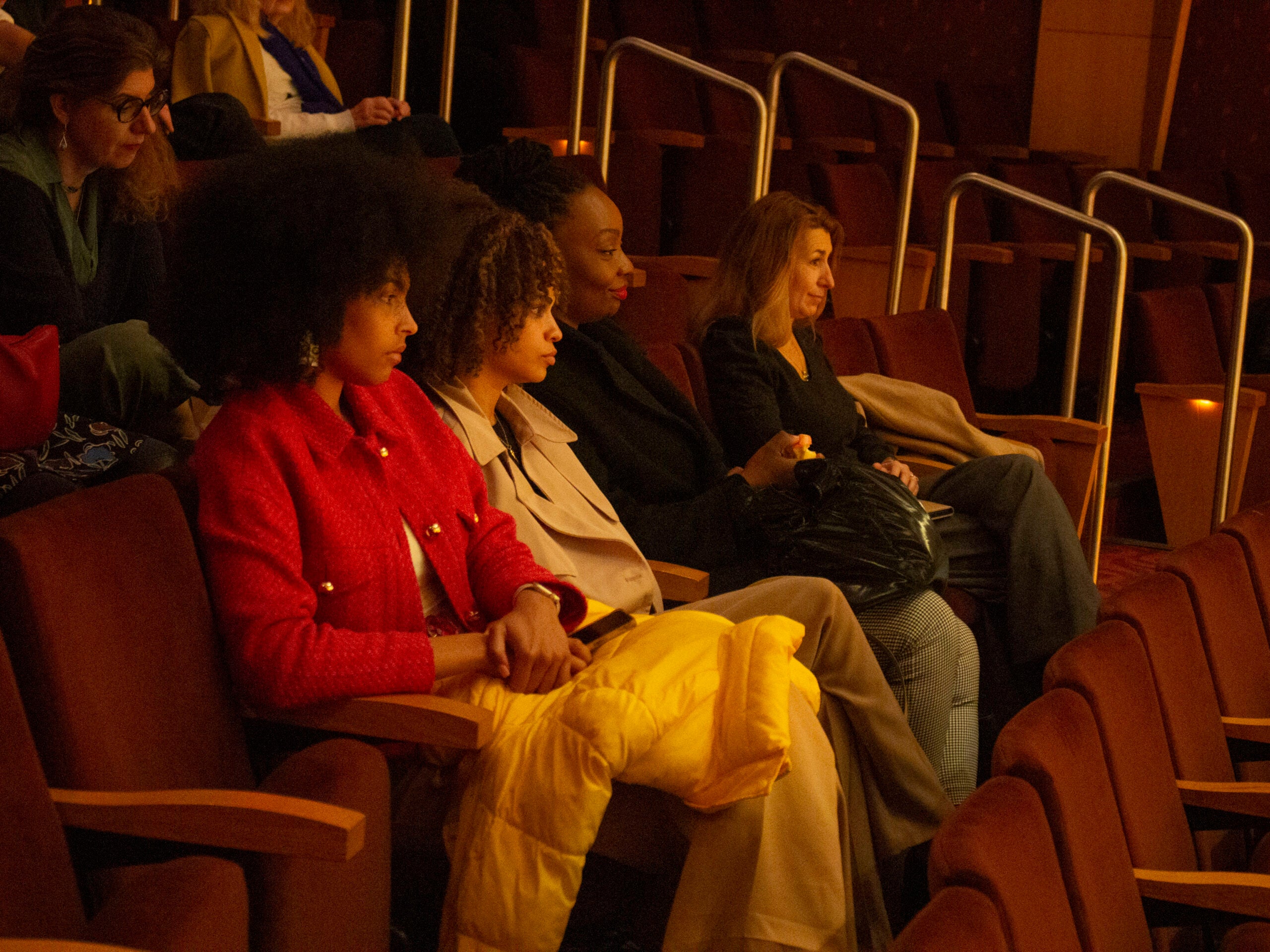 Four women are seated together in a lighted auditorium. They are watching the Q+A discussion.