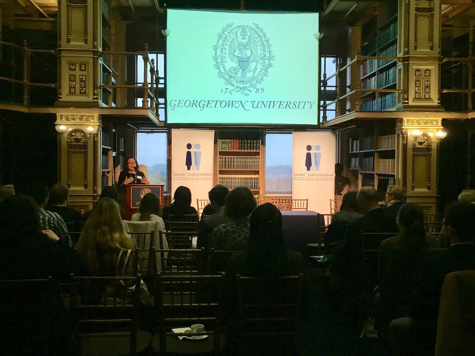 A seated audience listening to a woman speaking at a podium in a large library.