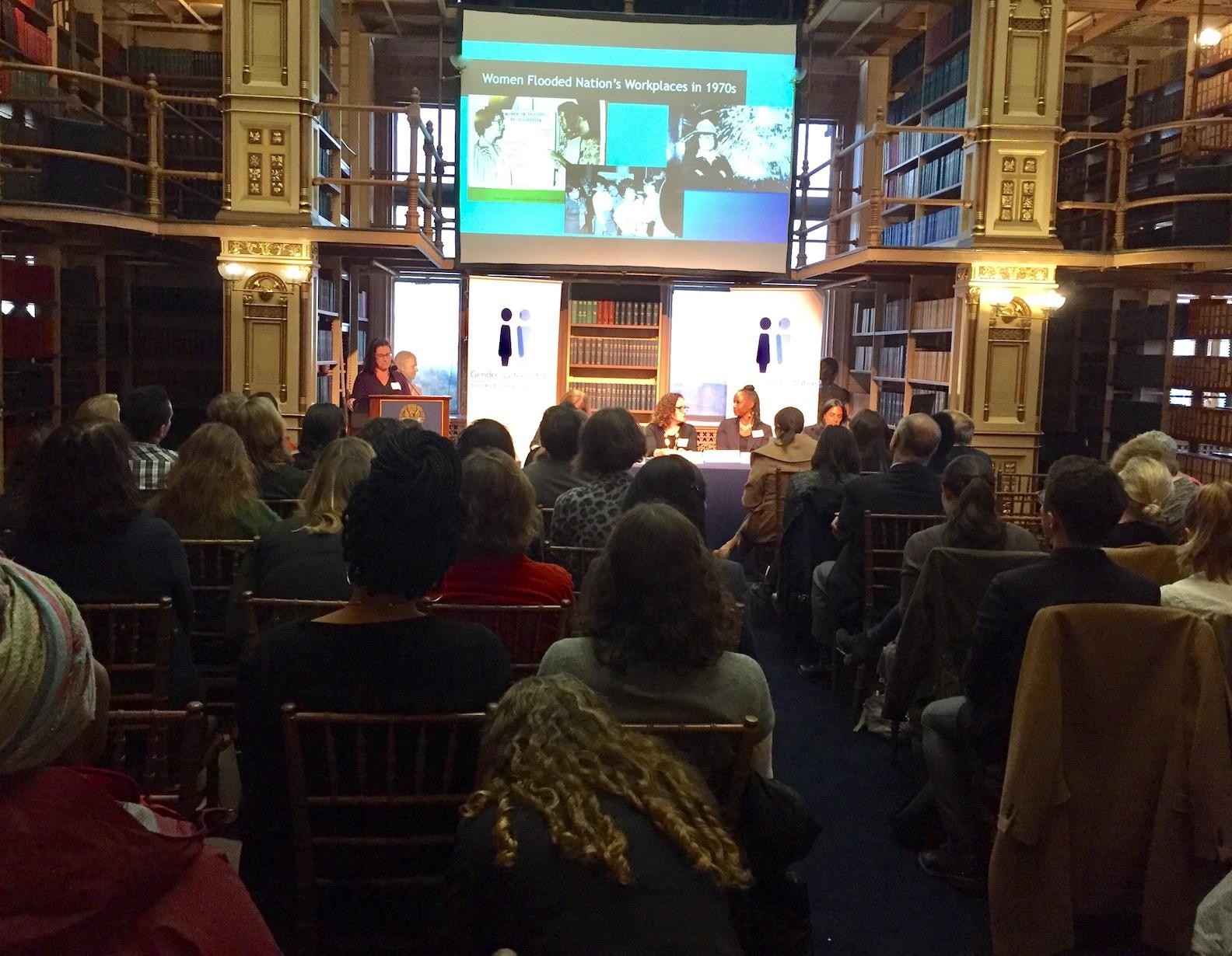 A seated audience listening to a woman speaking at a podium in a large library.