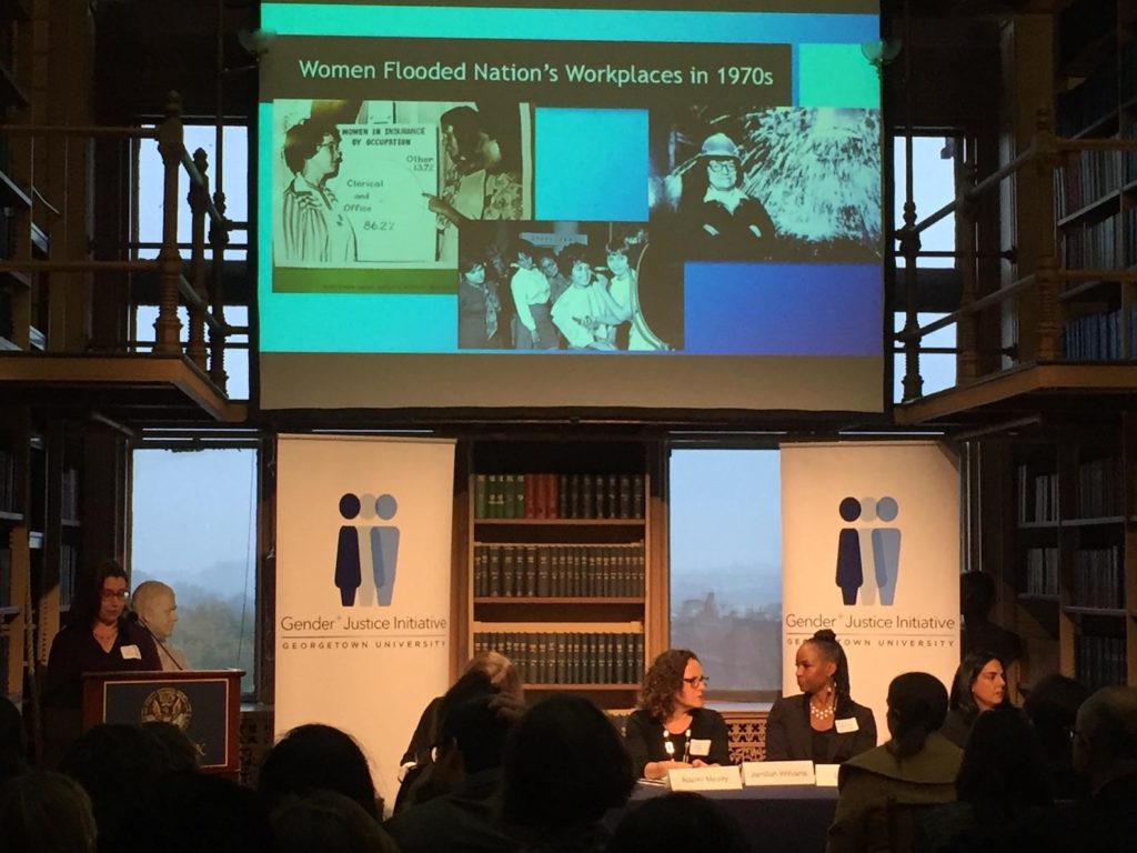 Three women sitting at a table for a panel discussion with a presentation behind them. They are speaking with eachother.