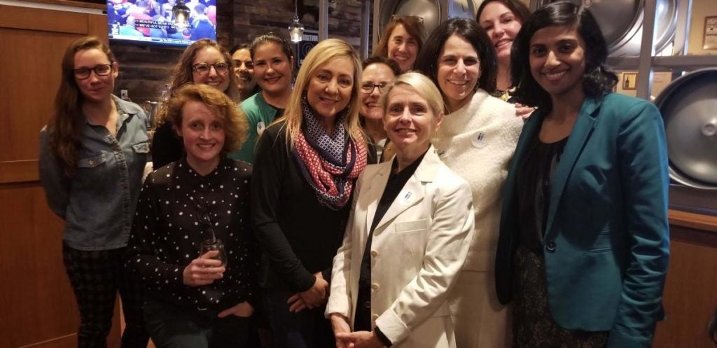 Several women stand and pose for a photo. Behind them is a bar with a tv mounted on the wall.