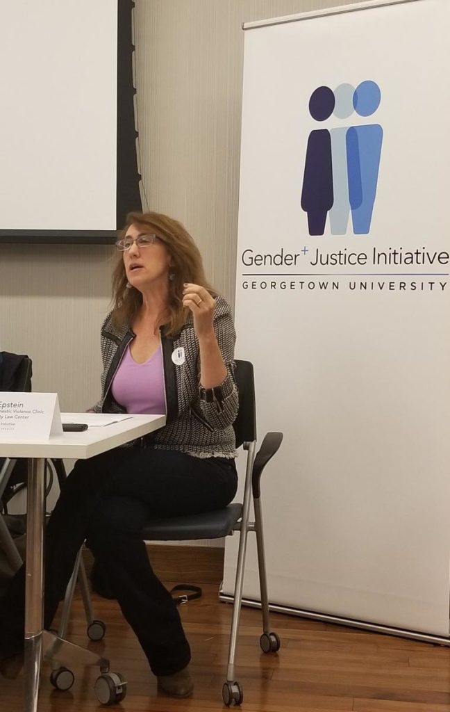 A woman sits in a chair behind a table. Behind her is a sign with the Gender+ Justice Initiative logo.