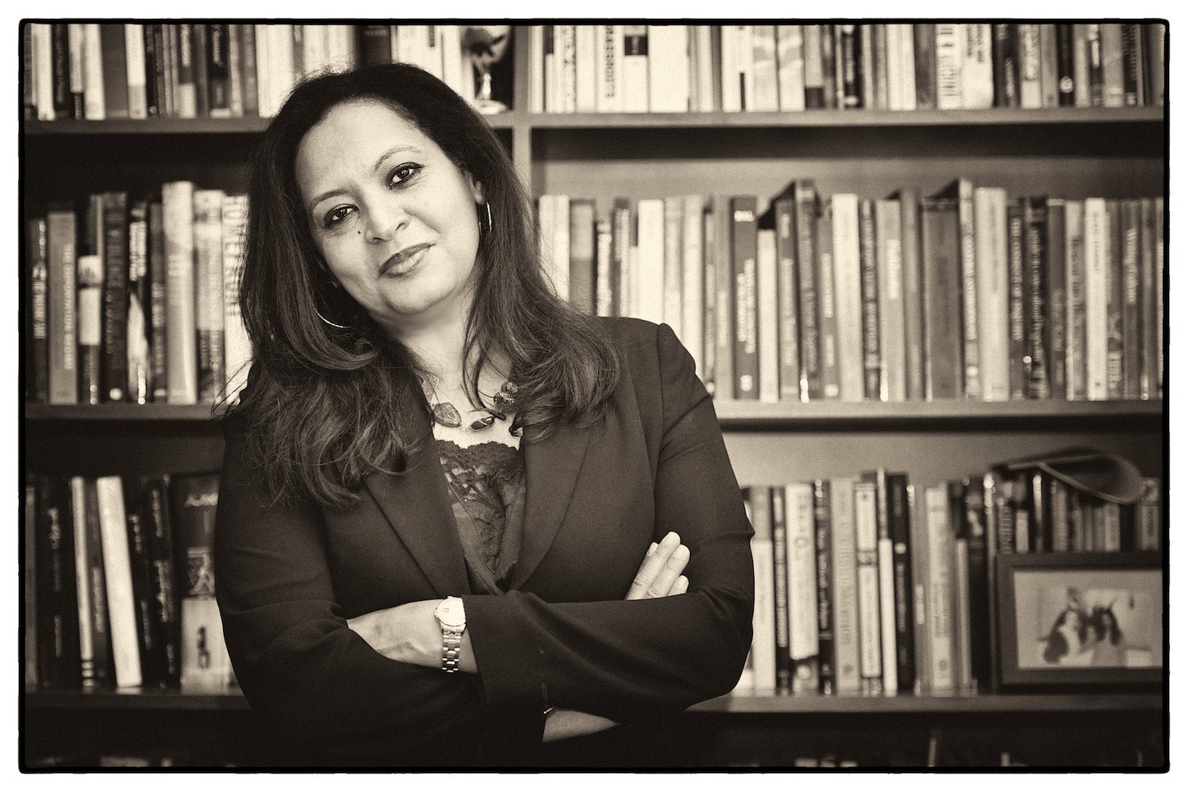 A woman with shoulder length hair wearing a blazer. Her arms are crossed and she is tilting her head to the left, smiling slightly. Behind her are several shelves of books.