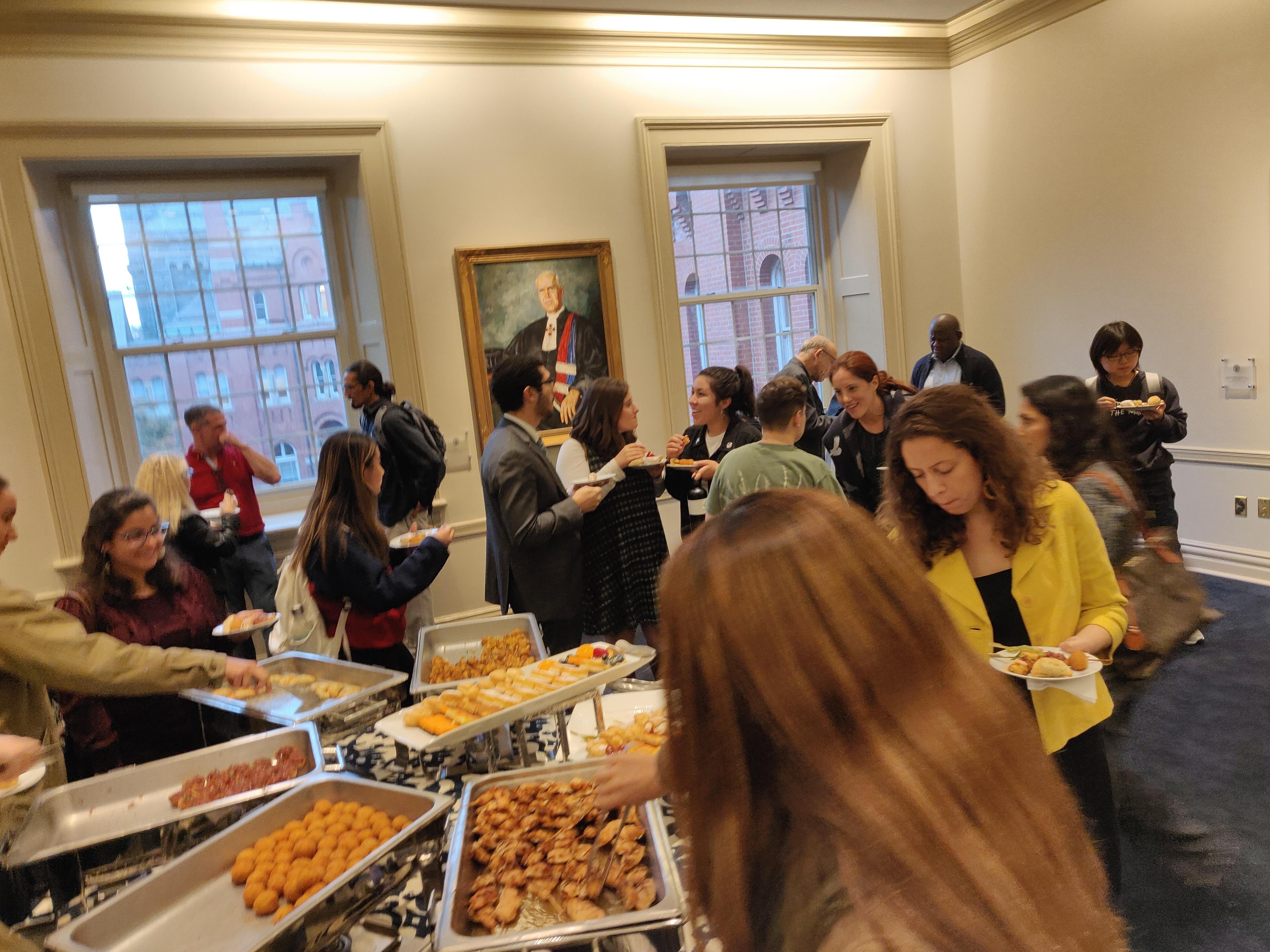 People lined up around a table of food, adding food to their plates.