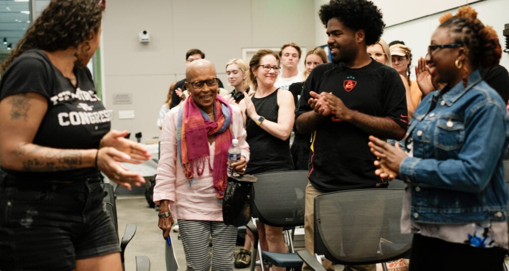 Dr. Patricia McFadden, an older, bald black woman wearing a pink shirt and scarf, is walking down an aisle. People applaud her in a standing ovation.