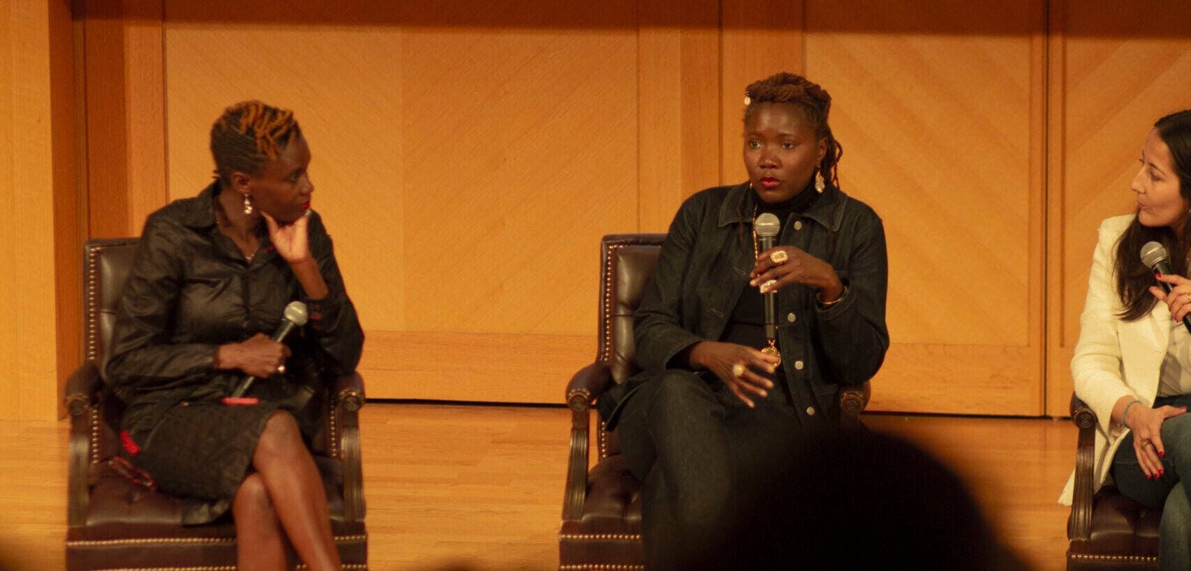 Three women seated on stage. The woman in the middle is speaking into a microphone, and the others look at her. Behind them is a screen projecting the event flyer for the movie screening of Saint Omer.