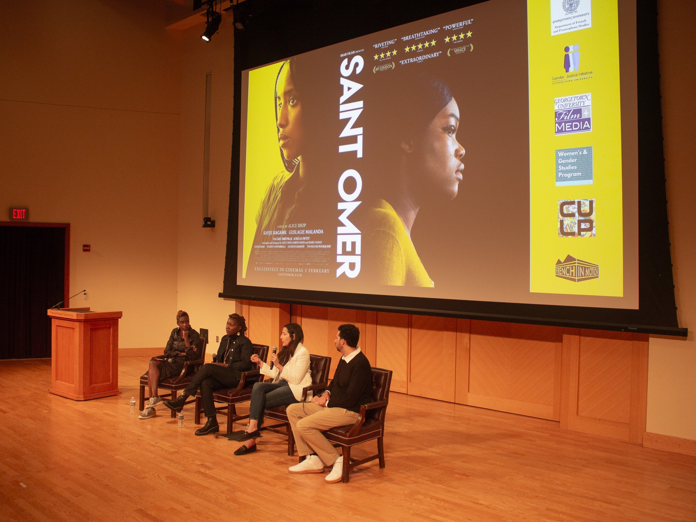 Four people sitting on a stage next to a podium. Behind them is a screen projecting the event flyer for the movie screening of Saint Omer.