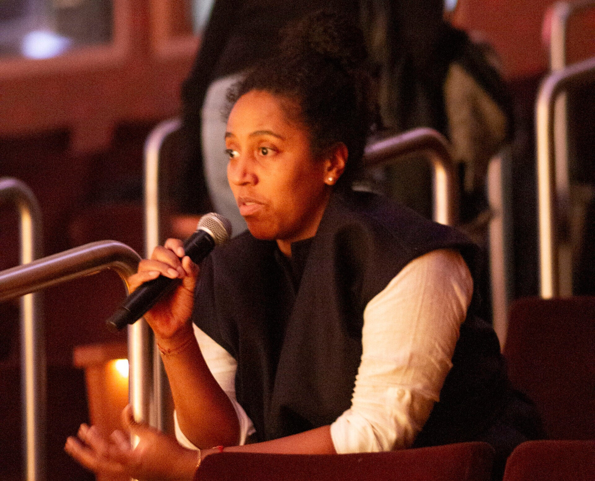 A woman in the audience speaking into a microphone, leaning forward and gesturing with her left hand.