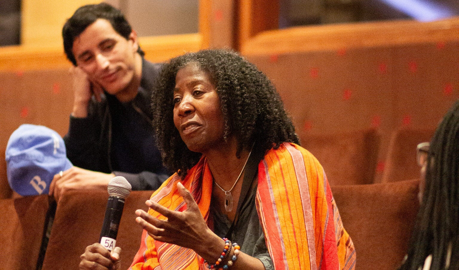 A woman sitting in the audience, speaking into a microphone. A man in the row behind looks at her, head in hand.