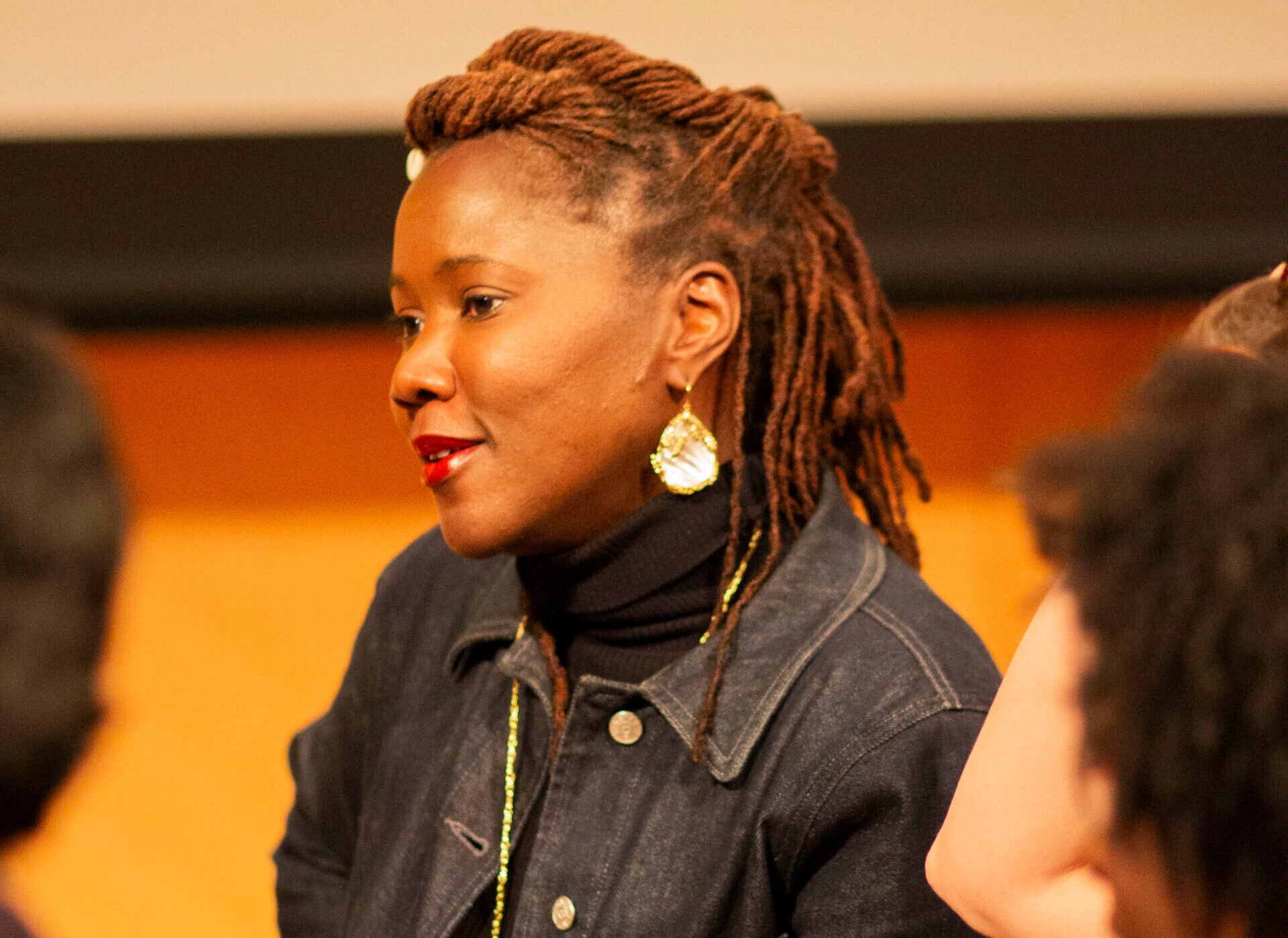 The side profile of a black woman with long hair in a tight twist style, wearing a blue jean jacket and gold jewelry.