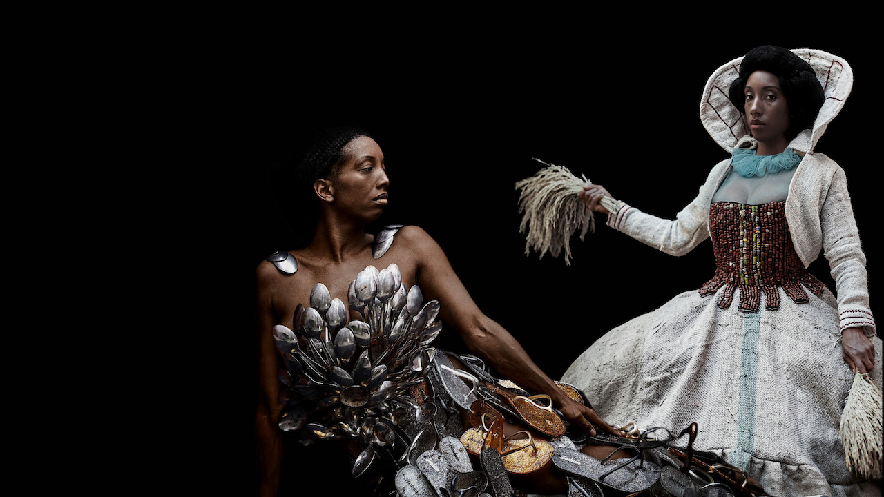 Artistic picture. Black background, left of image Black person lying on the side wearing a dress made of silver spoons and glittery flip flops, right of image, Black person wearing a white dress with a large ruff and a corset made of brown beads.