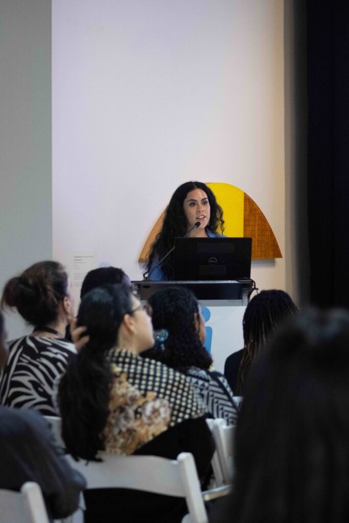 A woman standing behind a podium with a microphone. She is speaking and looking to the right.