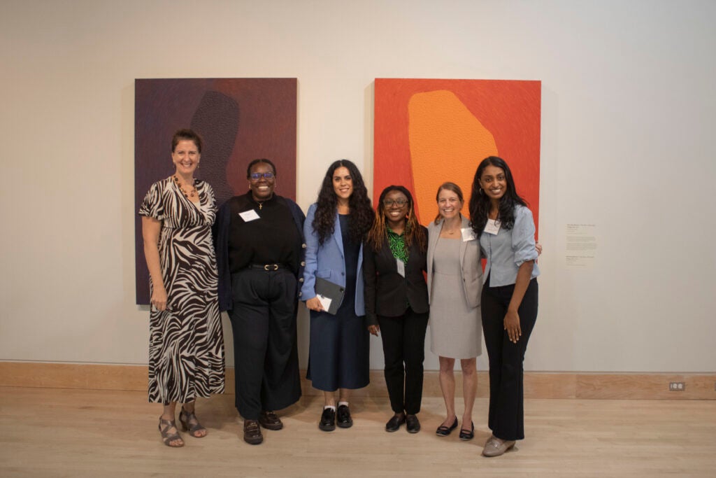 Six women standing in a line and smiling. Behind them are two large purple and orange pieces of artwork hanging on the wall. From left to right: Heather Bradford, Sherie Gayle, Paulina Macias Ortega, Halimat Somotan, Myka Held, Kirtana Ananth.