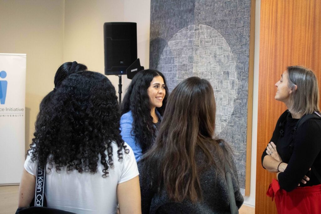 Five women standing in a group. Two womens' faces are visible, and they smile while talking to each other.