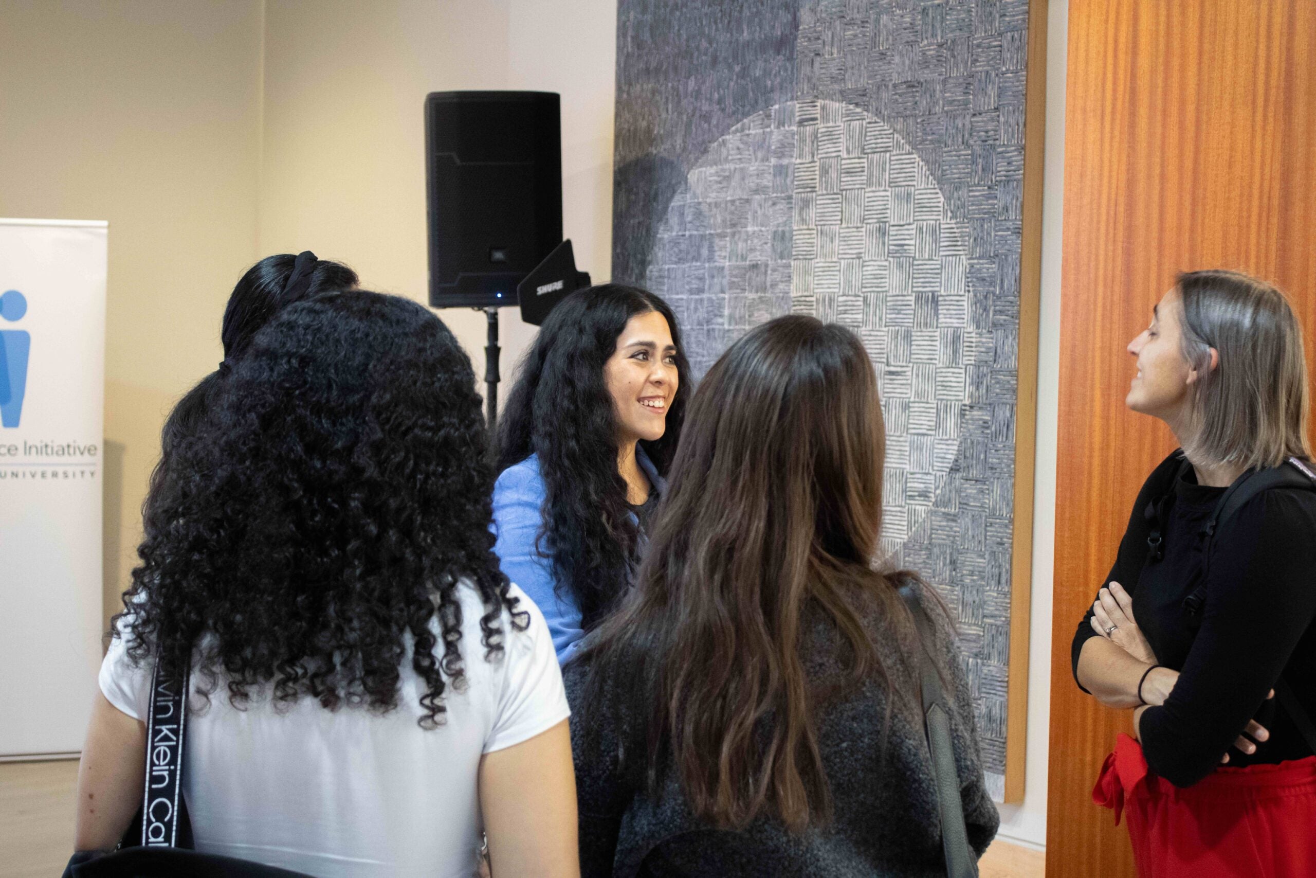 Five women standing in a group. Two womens' faces are visible, and they smile while talking to each other.
