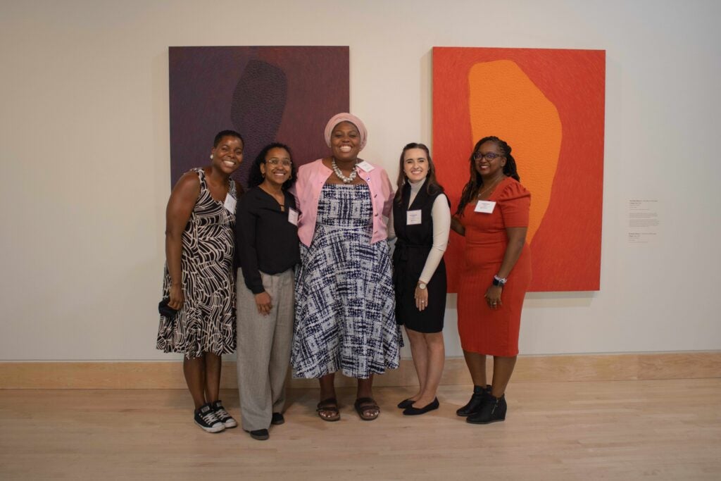 Five women standing in a line and smiling. Behind them are two large purple and orange pieces of artwork hanging on the wall. From left to right: DeJa Love, Alyssa Newman, Tuesday Barnes, Rachel Hanebutt, Amittia Parker.