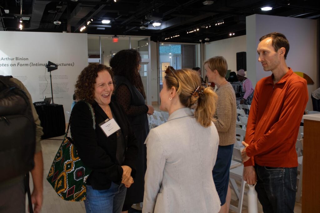 Two women having a discussion among a crowd of other people. The woman on the left is wearing a white name tag and is laughing. A man stands to the right, looking towards them.