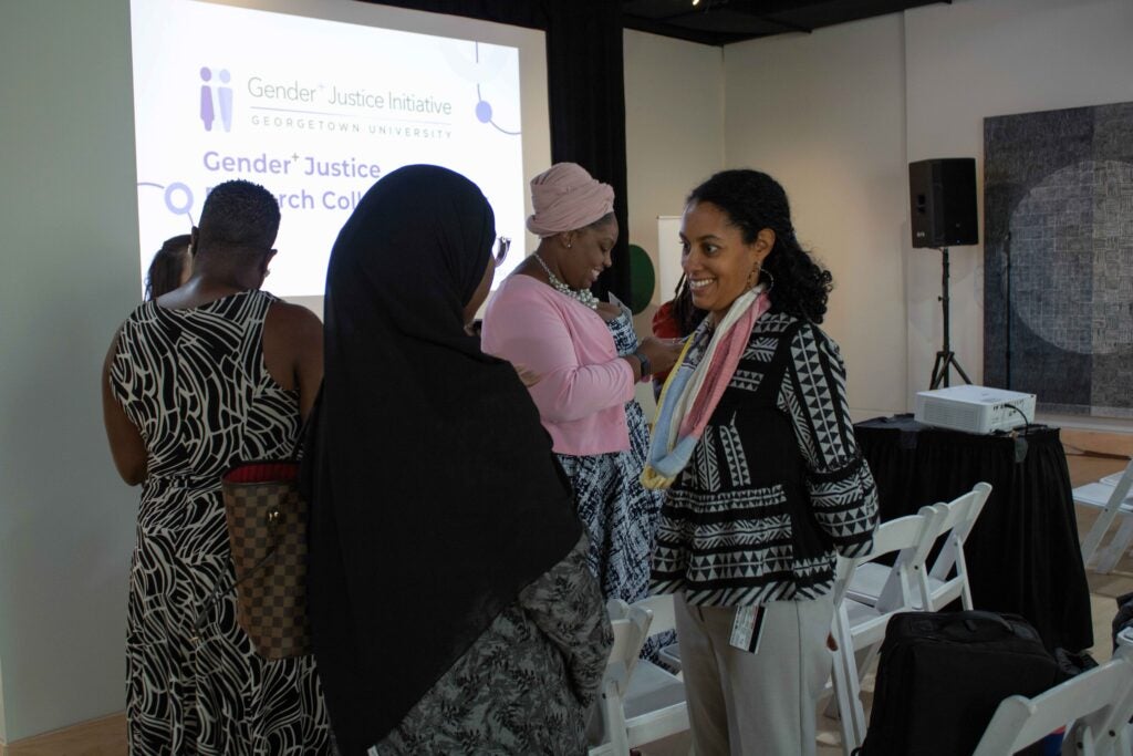 Two women standing and talking among other workshop participants. The woman on the right is smiling with her hands behind her back, and the woman on the left has her back turned to the camera.