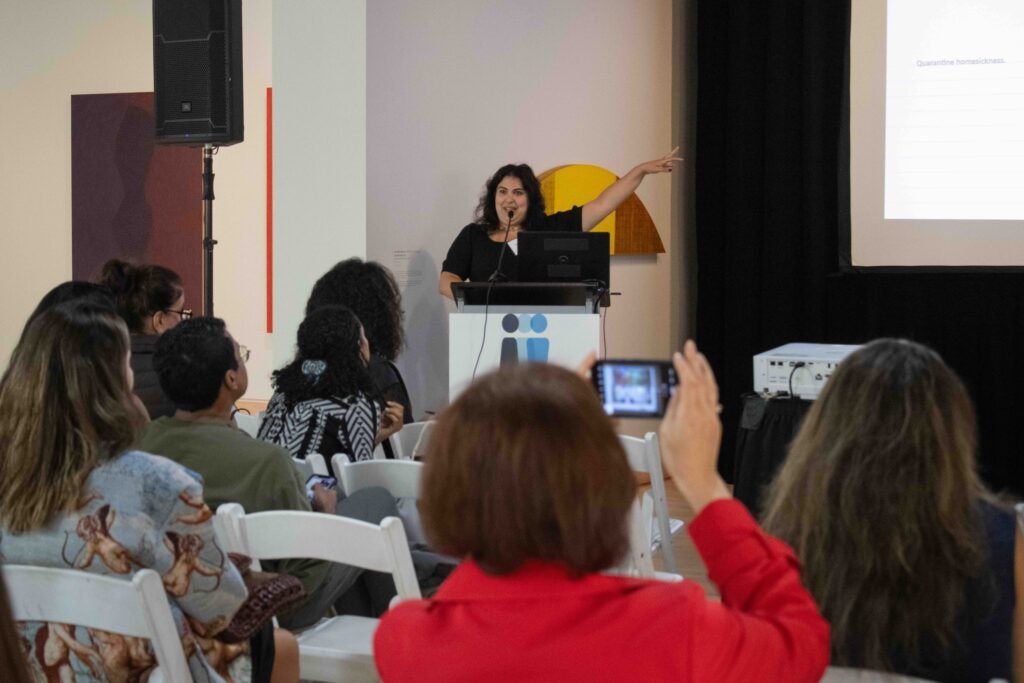 A woman standing behind a podium with a microphone. She is speaking and gesturing a hand up in the air to the right.