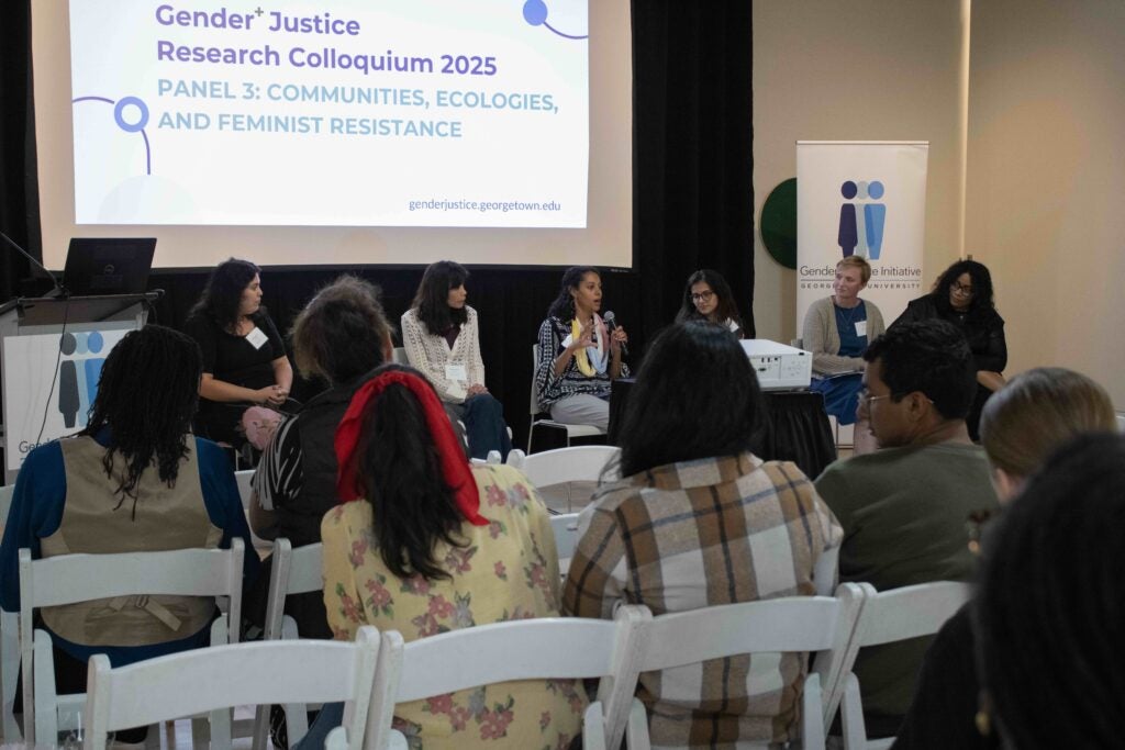 Six panelists sit in front of a projector with a white screen reading "Gender+ Justice Research Colloquium 2025. Panel 3: Communities, Ecologies, and Feminist Resistance." The woman in the middle is holding a microphone, and the other panelists look towards her.