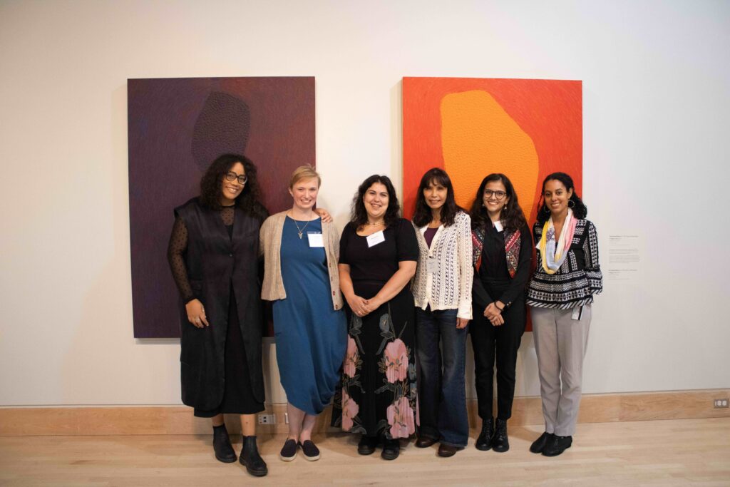 Six women standing in a line and smiling. Behind them are two large purple and orange pieces of artwork hanging on the wall. From left to right: Jennifer Boum Make, Erin Twohig, Heba Ghannam, Martina Thorne, Syeda Haleema Hasan, Lauren Arrington.