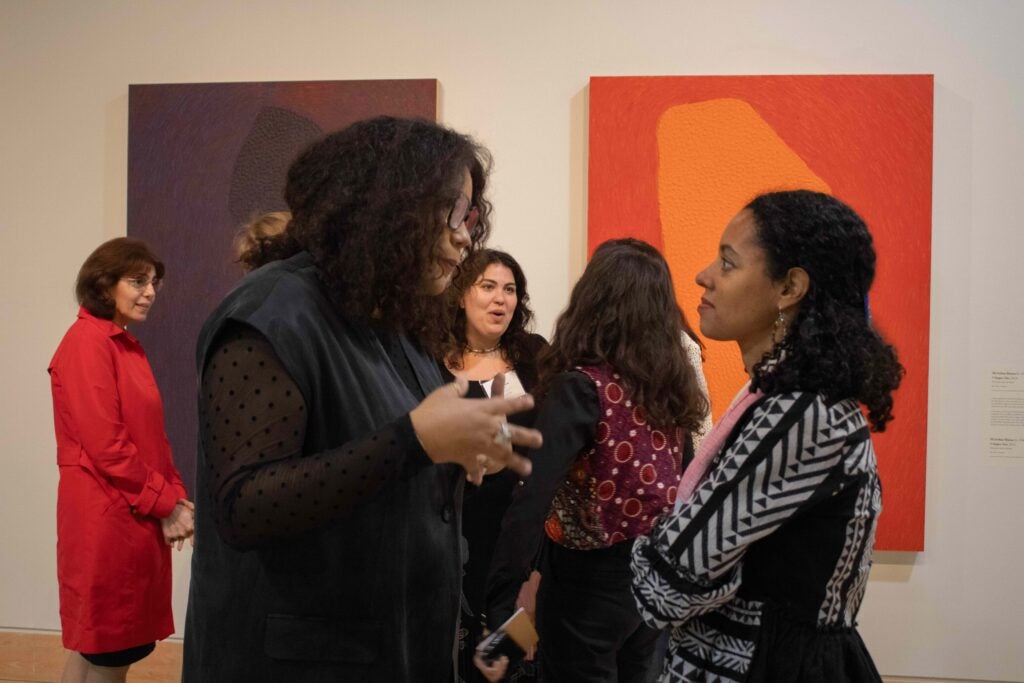 Two woman having a conversation among a crowd. The woman on the left gestures with her hands in front of her. The woman on the right has her arms crossed in front of her.