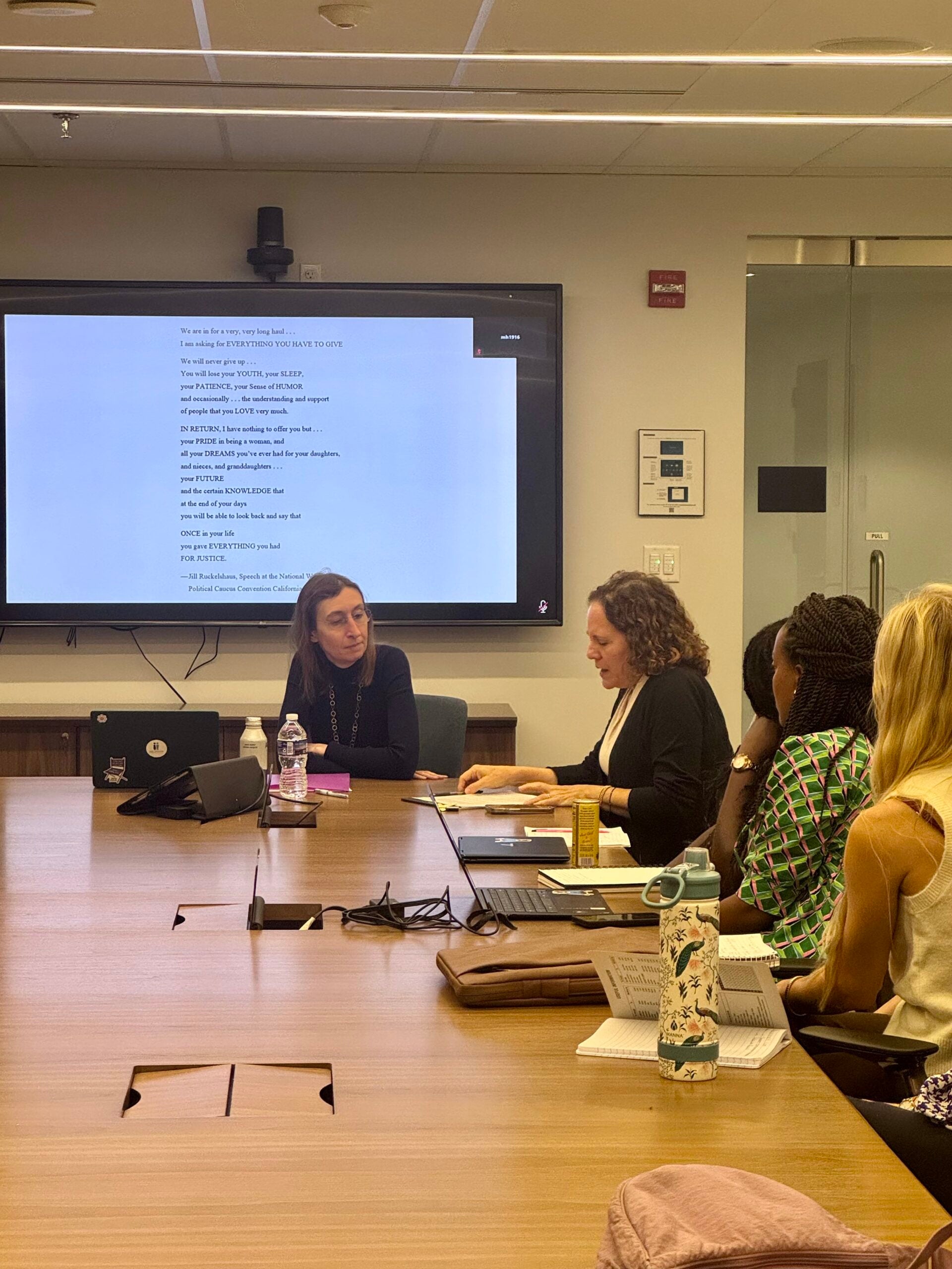 People sitting around a conference table. Behing the woman at the head of the table is a tv.