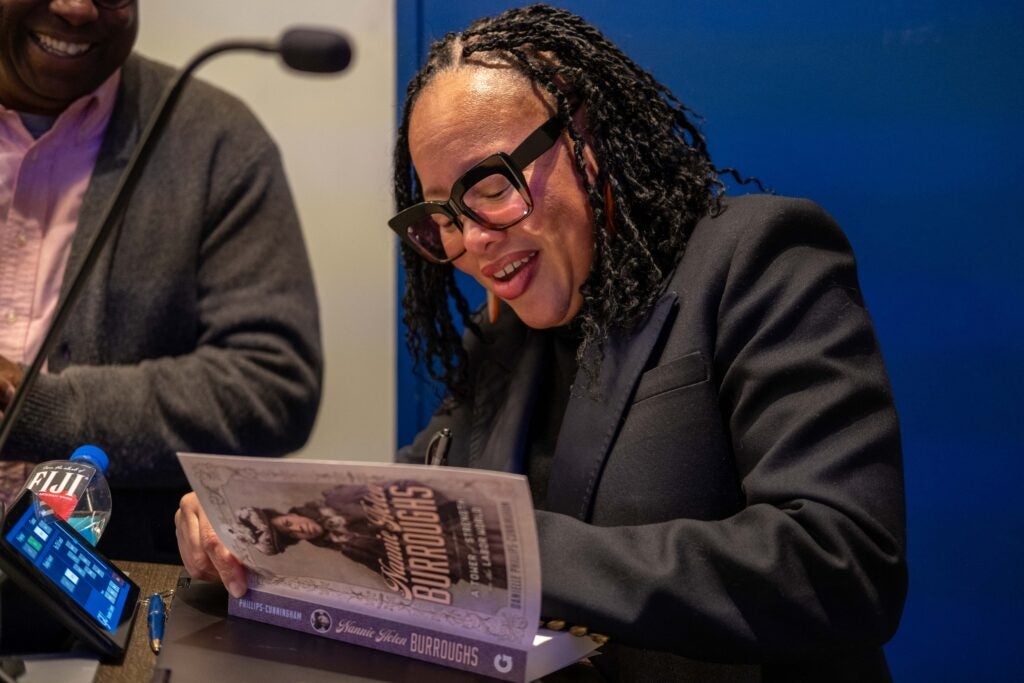 A woman wearing a black suit jacket smiling and signing the inside of a book titled “Nannie Helen Burroughs.” She is smiling, and a man standing behind her to the left smiles while watching.