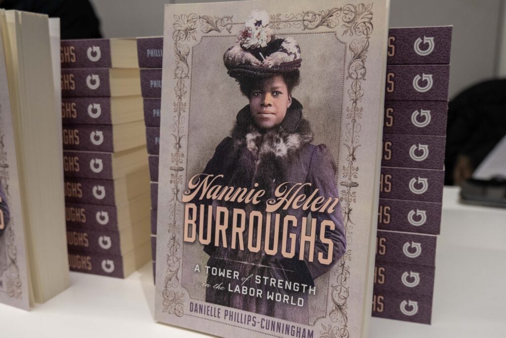 A stack of books with a purple spine. Another copy of the book is leaned upright against the stack. It features a woman in a large purple coat and the title reads “Nannie Helen Burroughs: a Tower of Strength in the Labor World” by Danielle Phillips-Cunningham.