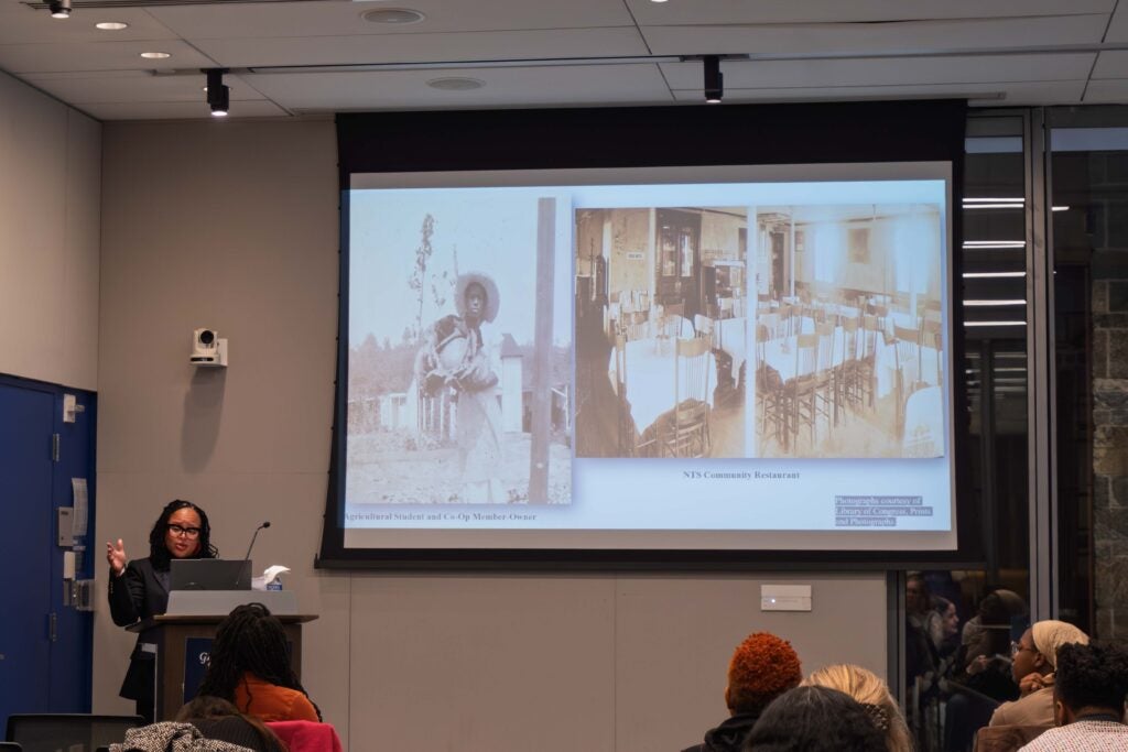 A woman stands behind a podium talking while gesturing a hand in the air. On the wall behind her is a large projector screen showing old photographs. Several people sitting in the audience watch.