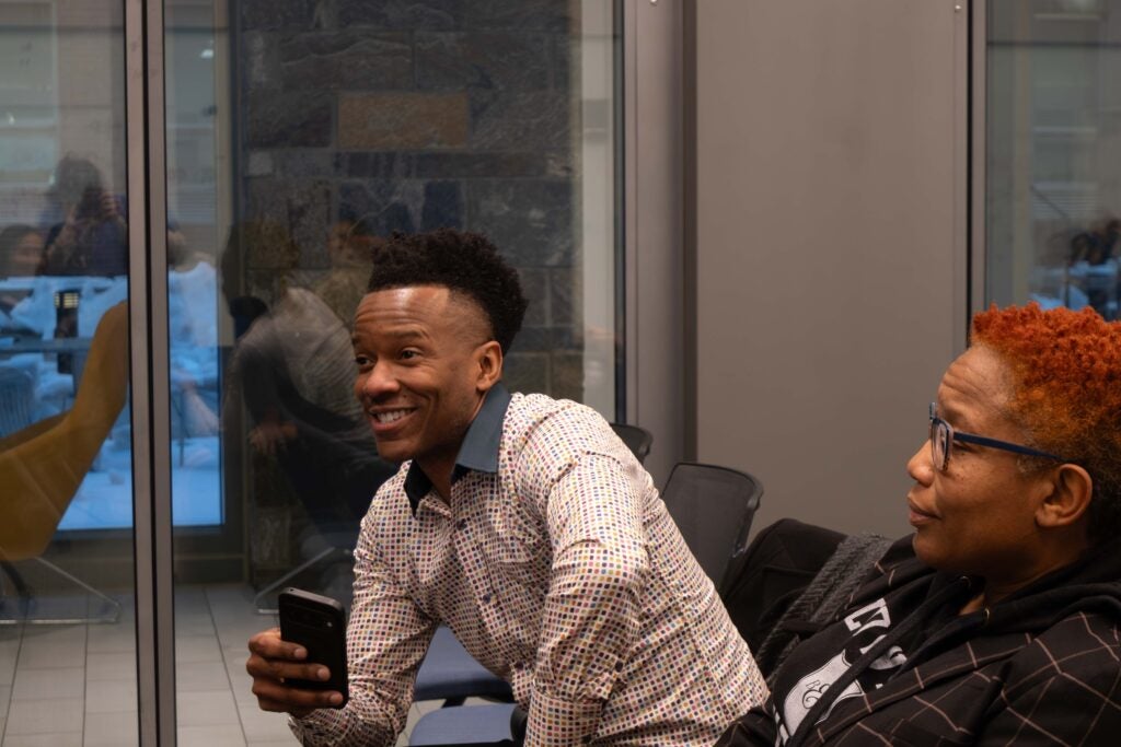 A man sitting in the audience smiles and leans forward with one hand on his knee and the other hand holding a cell phone in front of him. A woman sitting next to him sits back in her chair.