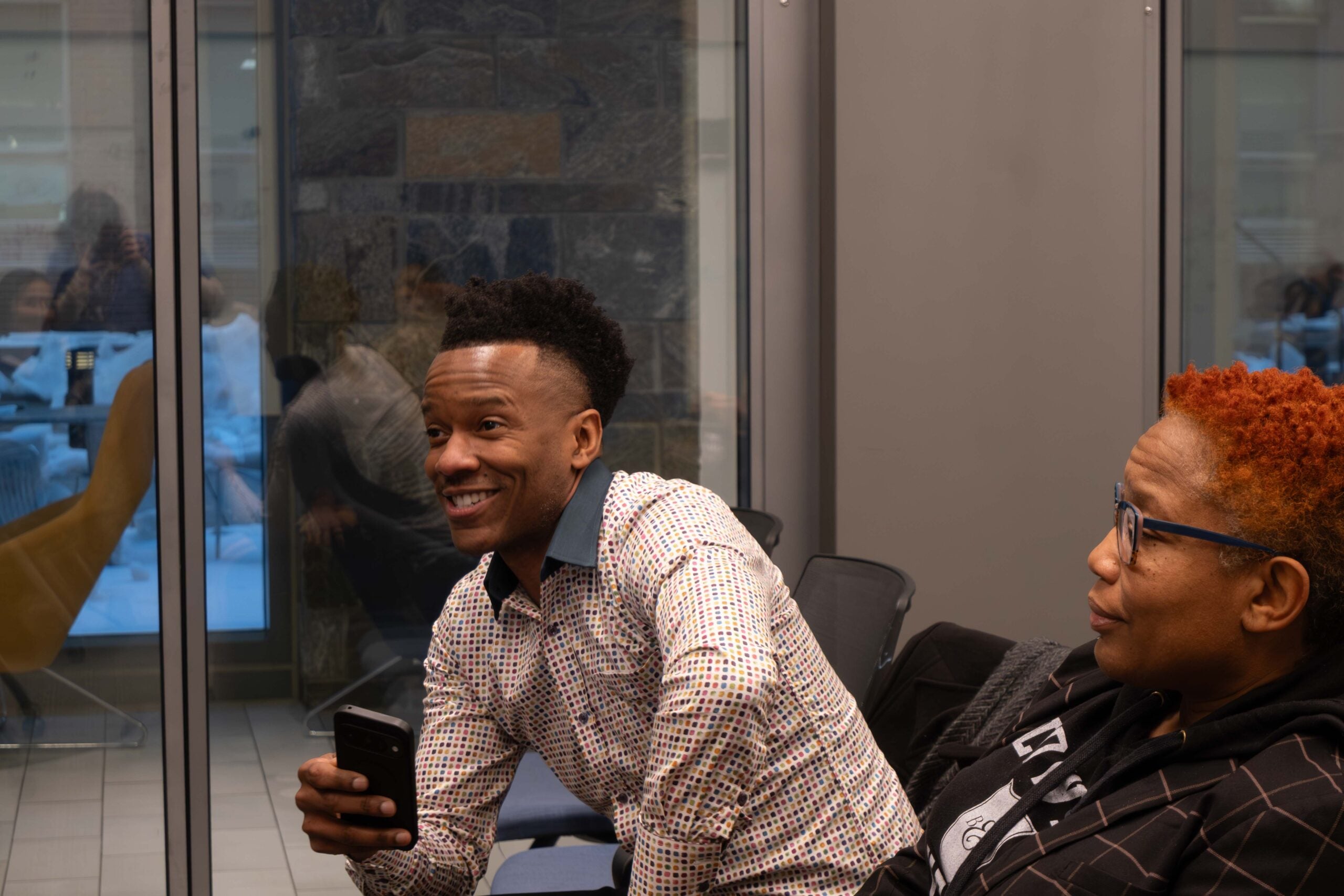 A man sitting in the audience smiles and leans forward with one hand on his knee and the other hand holding a cell phone in front of him. A woman sitting next to him sits back in her chair.