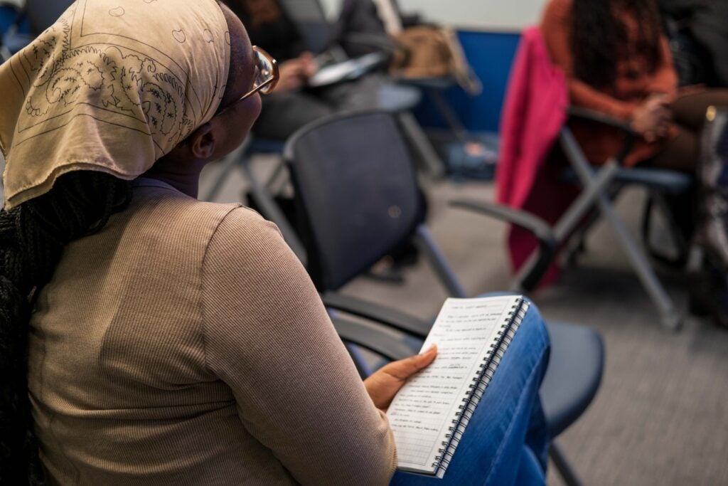 The back of a woman sitting with her legs crossed and head tilted slightly to the left. She is holding a notebook with notes written on it in her hand.