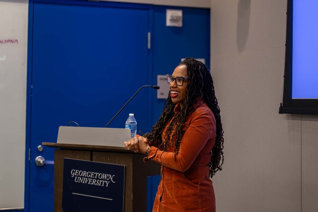 A woman with long dark hair smiles and stands next to a wooden podium with a sign on it reading “Georgetown University.” Her hands are clasped in front of her, resting on the podium.