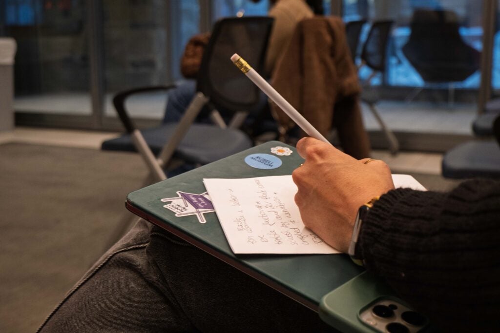 A close up photo of someone writing notes on a piece of paper with a pencil. The paper is resting on a laptop with a green cover and stickers in someone’s lap.