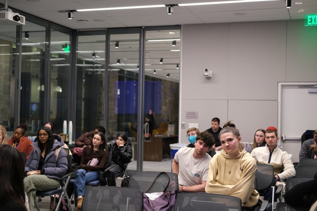 Audience members sitting in a room with glass walls, looking forward.