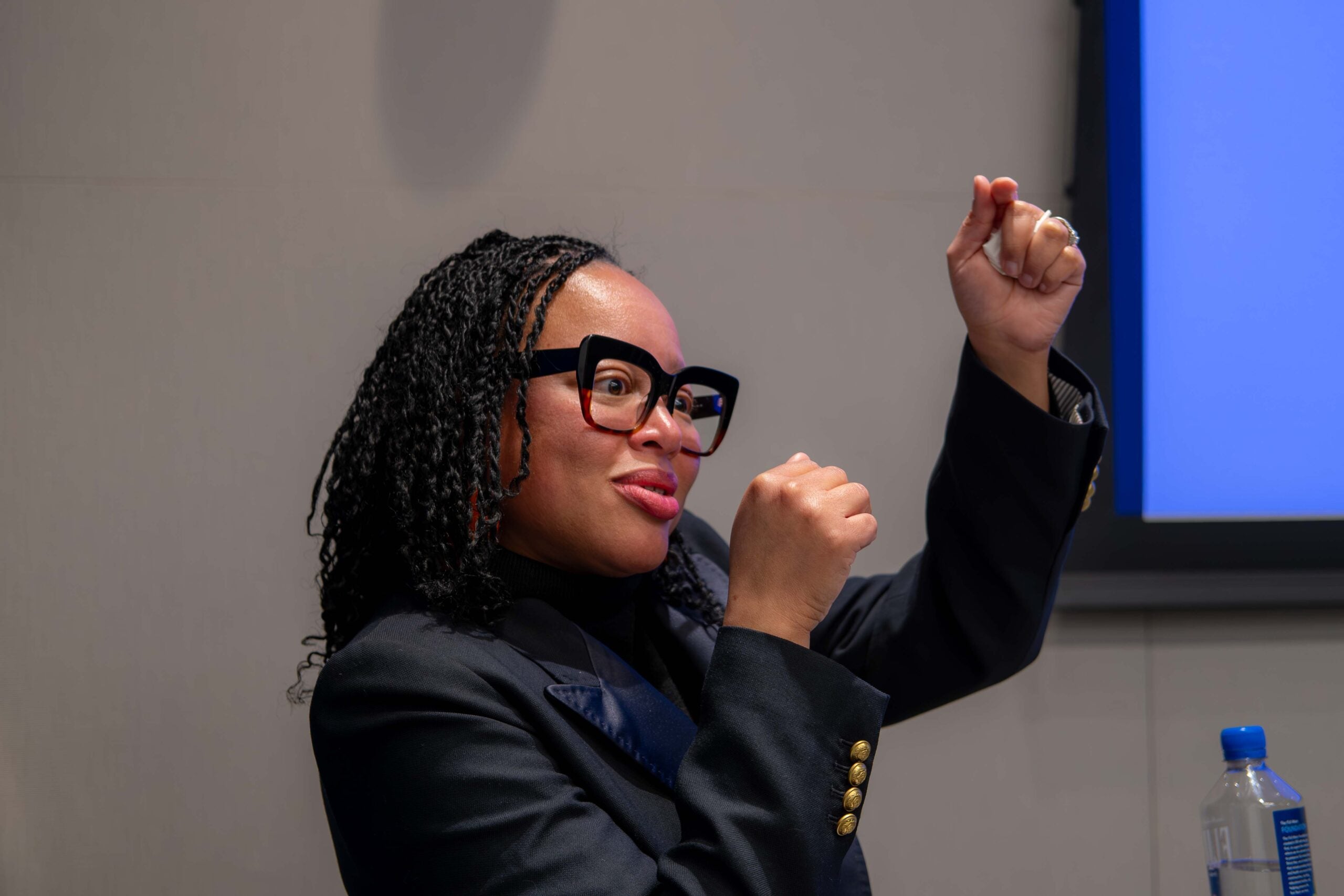 A woman in a black suit jacket standing behind a podium and gesturing in the air with both hands, looking to the right.