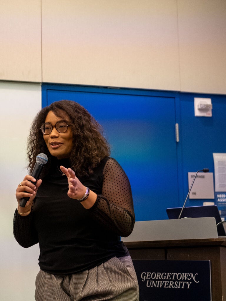 A woman with curly hair and large square rimmed glasses stands in front of a Georgetown University podium against a bright blue wall. She is holding a microphone with one hand and gesturing to the side with the other.