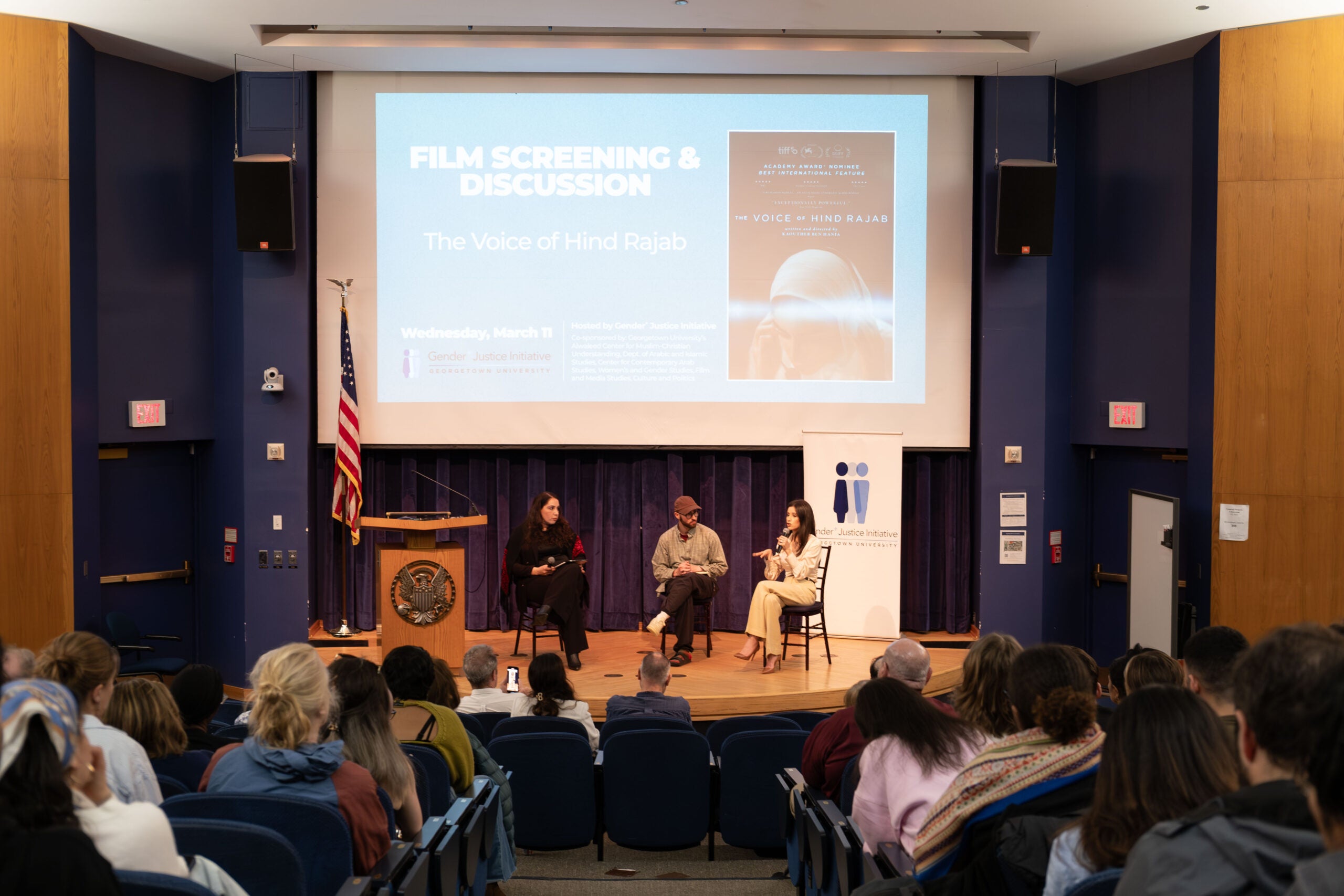 Three poeple sitting on a stage in front of an audience. The woman on the right is speaking into a microphone. Behind them a large projector screen displays the poster for the film The Voice of Hind Rajab.