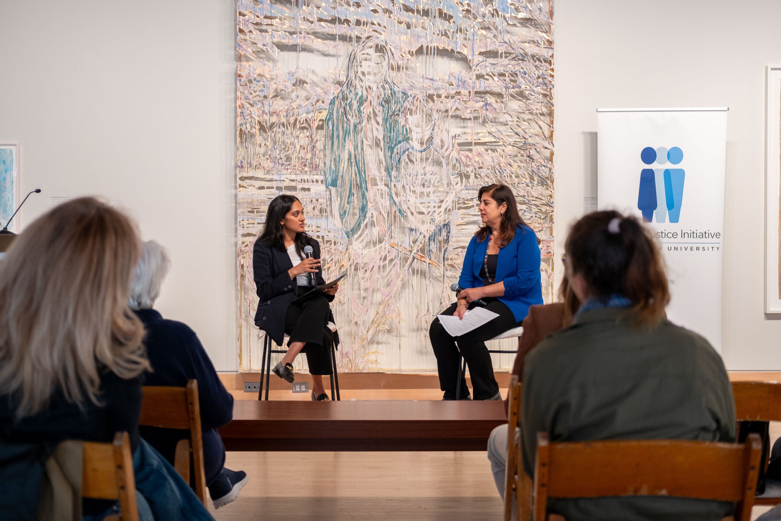 Two women sitting in chairs at the front of a room, facing each other. Artwork hangs from the wall behind them. An audience is seated on each side of an aisle, facing them.