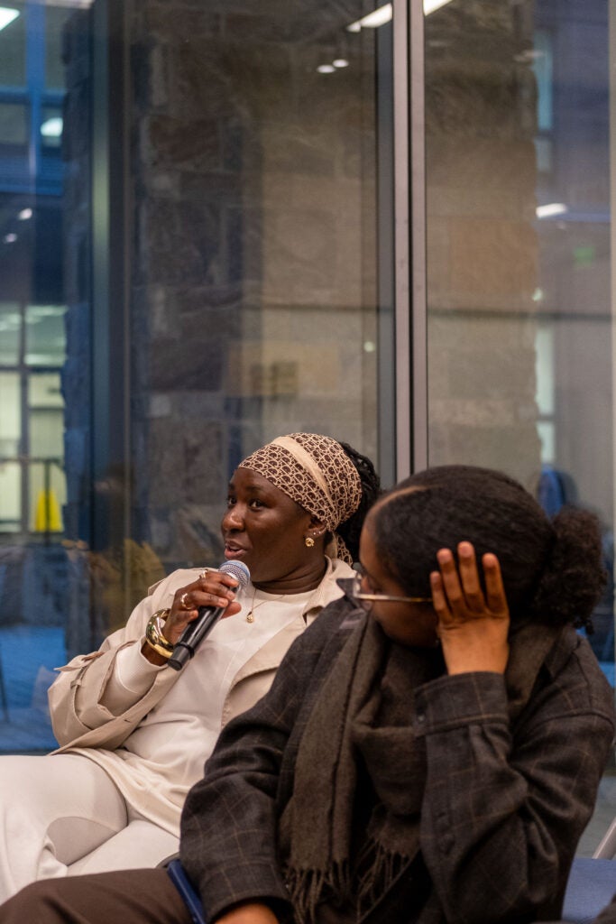 A woman from the audience sits and speaks into a microphone, looking forward. The woman on the right has her head propped up on her left hand and looks towards the speaking woman.