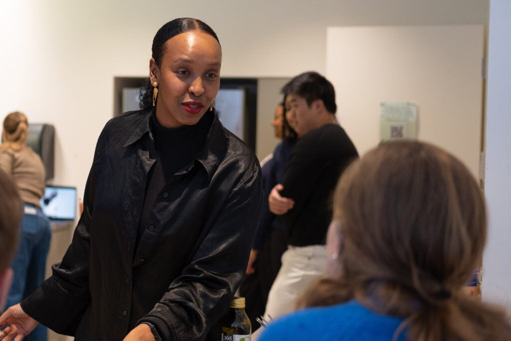 A woman in all black standing in a kitchen, talking to someone sitting on the other side of the counter.