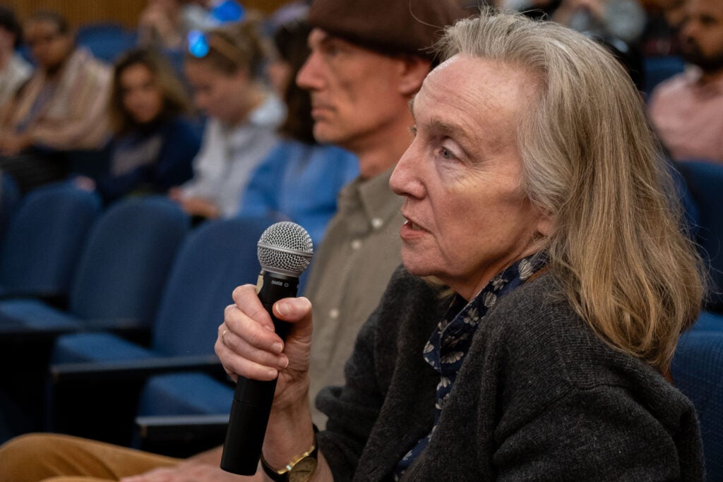 A close up side profile photo of a woman in the audience holding a microphone close to her face. She has medium length grey hair and is wearing a grey sweater over a collared button down shirt.