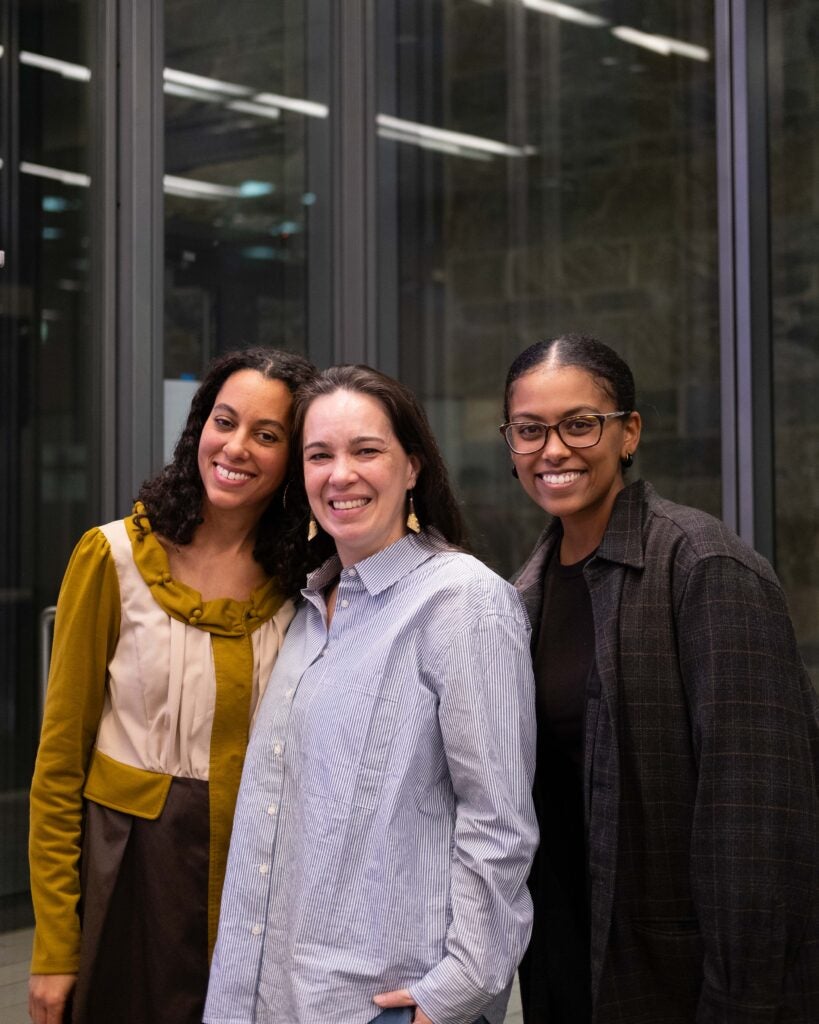 Three women smiling for a photo. The woman on the left is wearing a long sleeve yellow and beige shirt, the woman in the middle is wearing a blue and white striped button down shirt, and the woman on the right is wearing a grey and tan plaid button down shirt. 
