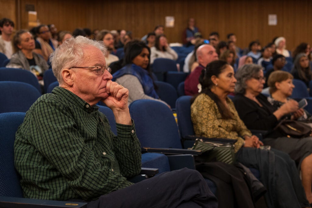 A man sitting in the audience with his legs crossed and head propped up in his hand. He is looking intently at the stage. Other audience members in the background also face the stage, out of view.
