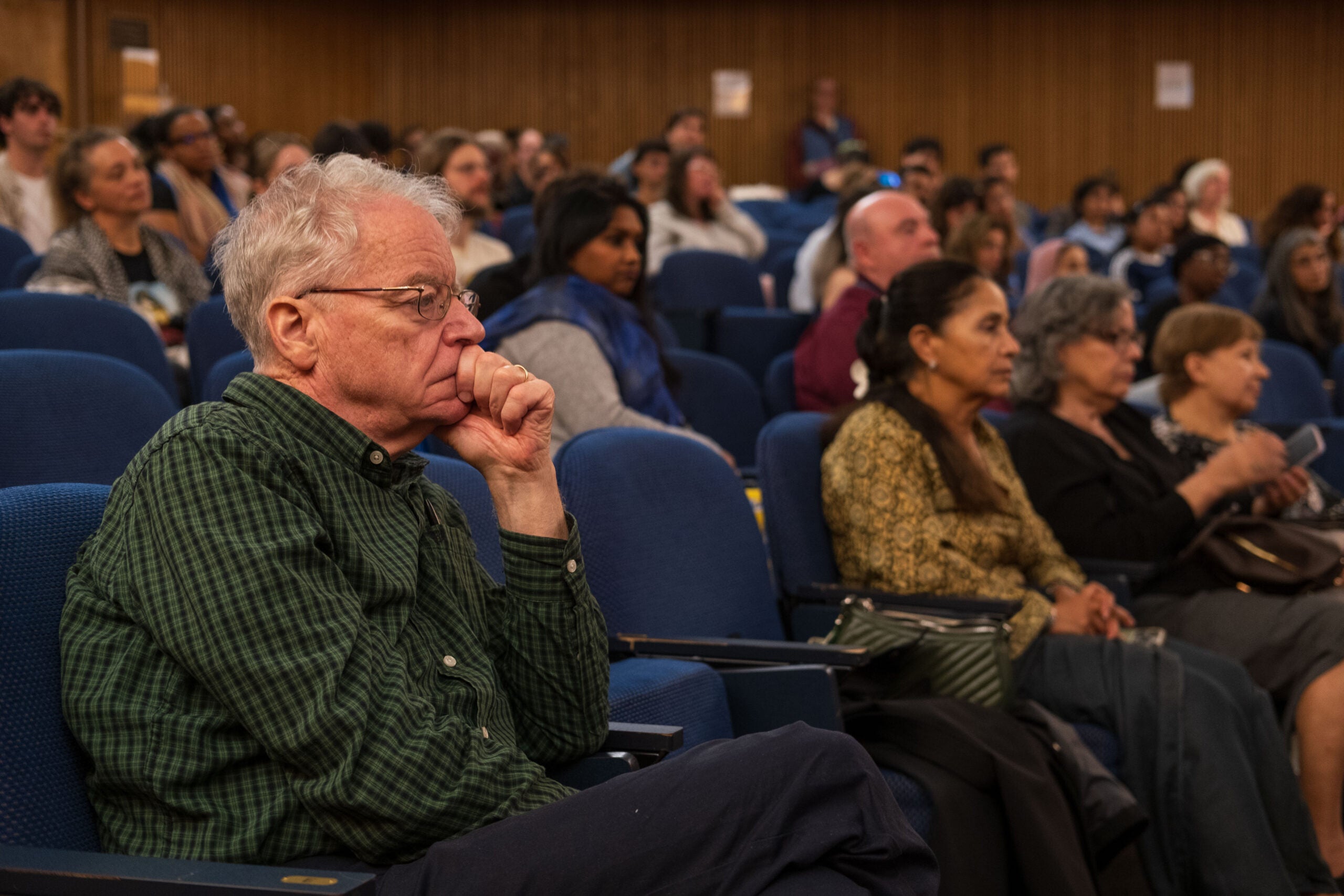 A man sitting in the audience with his legs crossed and head propped up in his hand, looking intently toward the stage out of frame.