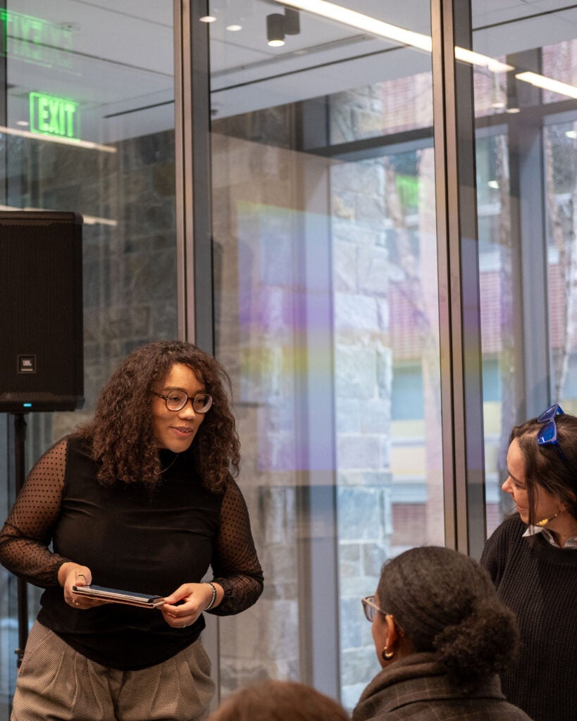 A woman with black rimmed glasses stands and leans slightly over to the right, talking to two audience members. She is smiling and holding an iPad in her hands.