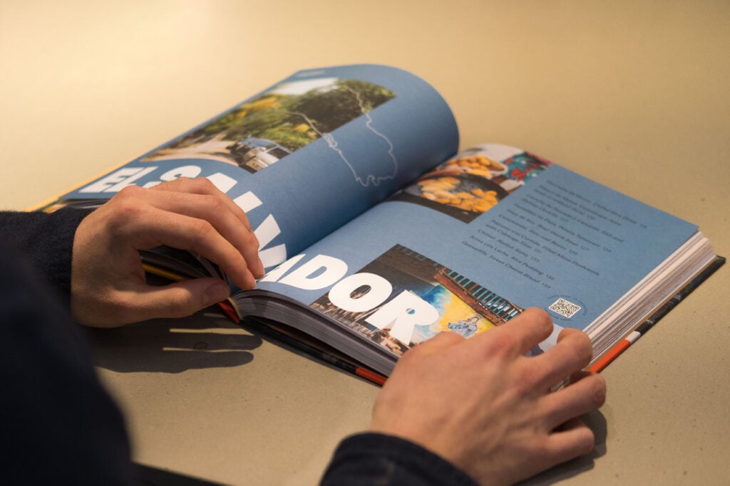 A pair of hands flips through a book. The open pages are blue and a large title at the bottom reads “El Salvador.”