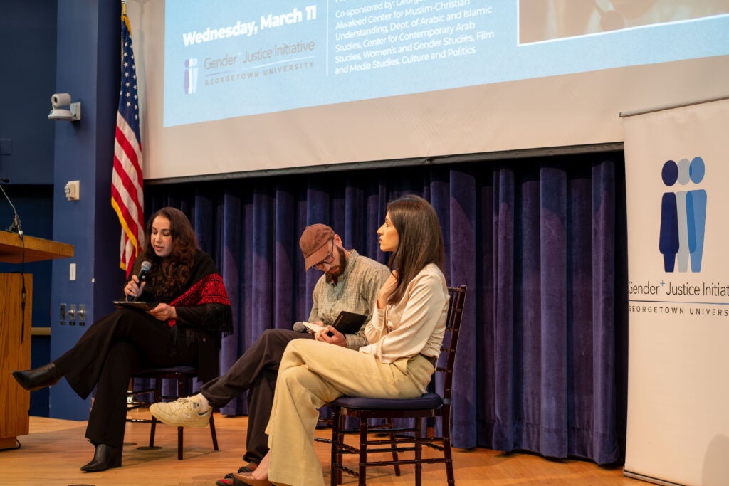 A close up side profile photo of two women and one man sitting on a stage. The woman on the left is speaking into a microphone. The man in the middle is looking down at the open notebook in his lap, and the woman on the right is looking up towards the ceiling.